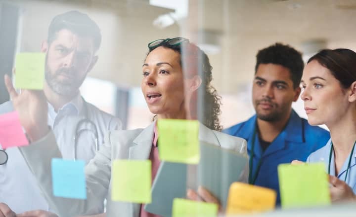 female healthcare leaders in a hospital setting reviewing healthcare supply chain management strategies with sticky notes on a glass wall